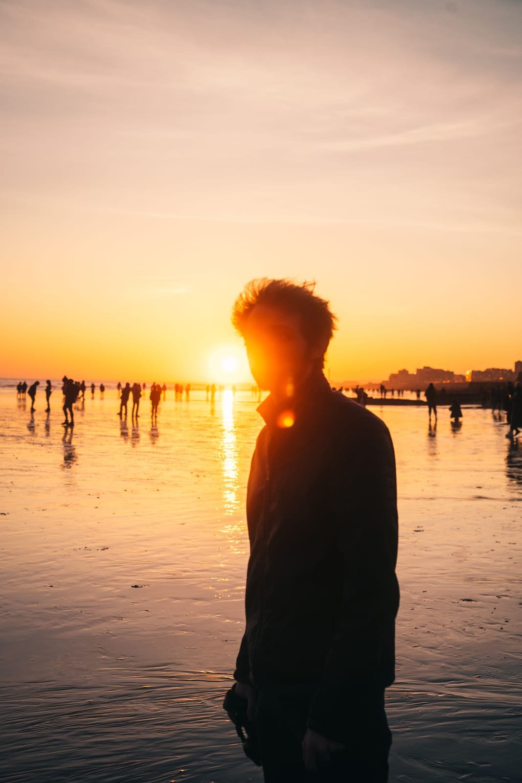 Silhouetted figure on a wet beach at sunset with a crowd in the distance