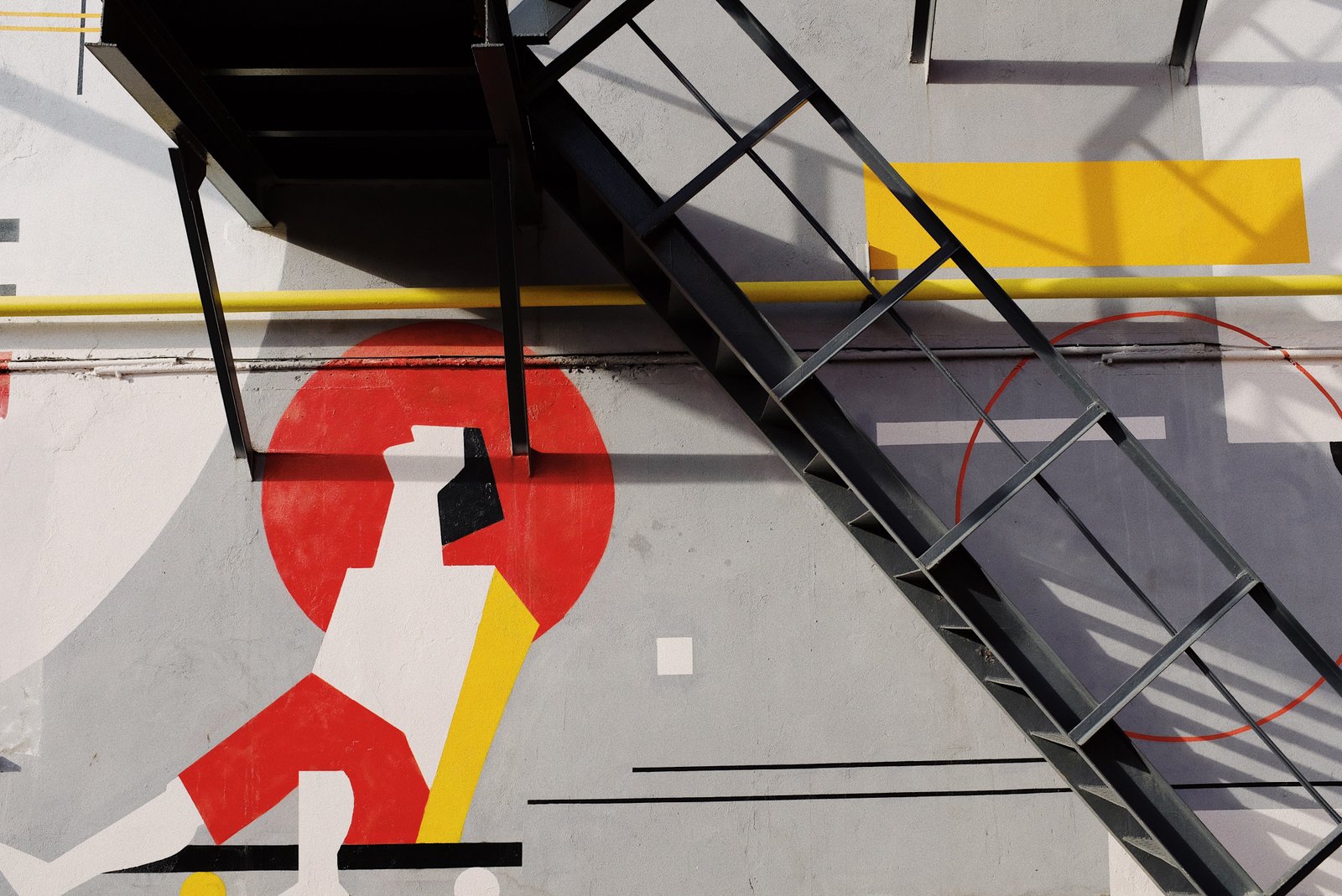 A diagonal fire-escape ladder cutting across a bold geometric mural, competing shapes and colors crowding the same wall